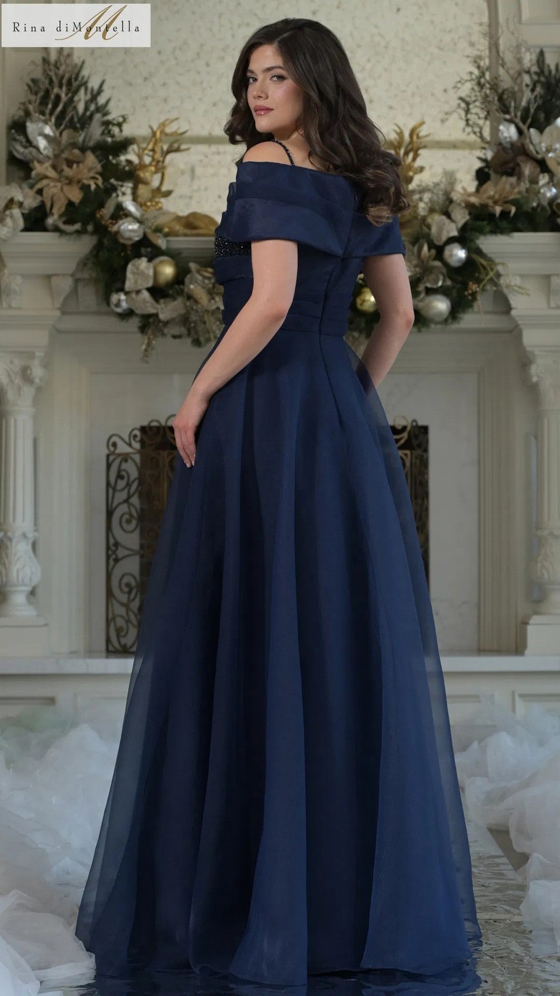 A woman in the Long Formal Dress: Rina Di Montella RD3303, a navy off-shoulder, floor-length gown, stands before a decorated white fireplace with floral arrangements.